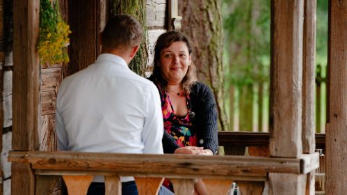 Basia siedzi na drewnianym ganku, naprzeciwko mężczyzny w jasnej koszuli. Kobieta ma na sobie kolorową sukienkę i ciemny sweter, a na jej twarzy widać delikatny uśmiech. Scena ma spokojny, sielski klimat, podkreślony naturalnym otoczeniem i ciepłym światłem.