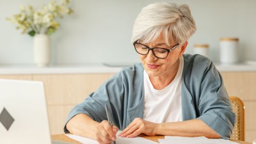 Starsza kobieta w krótkich siwych włosach i okularach z czarnymi oprawkami siedzi przed komputerem i ręcznie uzupełnia dokumenty leżące na stole.