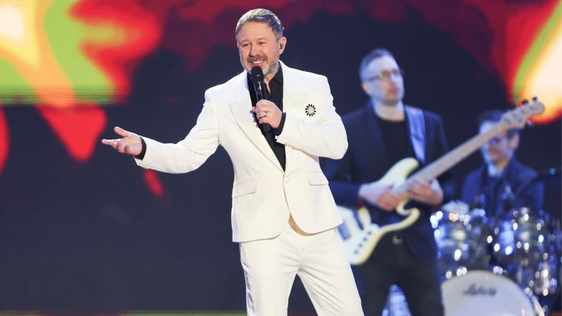 Andrzej Piaseczny śpiewa do mikrofonu. Jest ubrany w jasny garnitur i czarną koszulę.