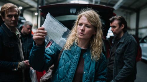 „Profilerka Jastrë” w TVP VOD. Julia Wigier w niebieskiej kurtce ogląda przezroczystą torebkę z dokumentem przy otwartym bagażniku samochodu. W tle trzy osoby i zaparkowane auta w przestronnej hali.