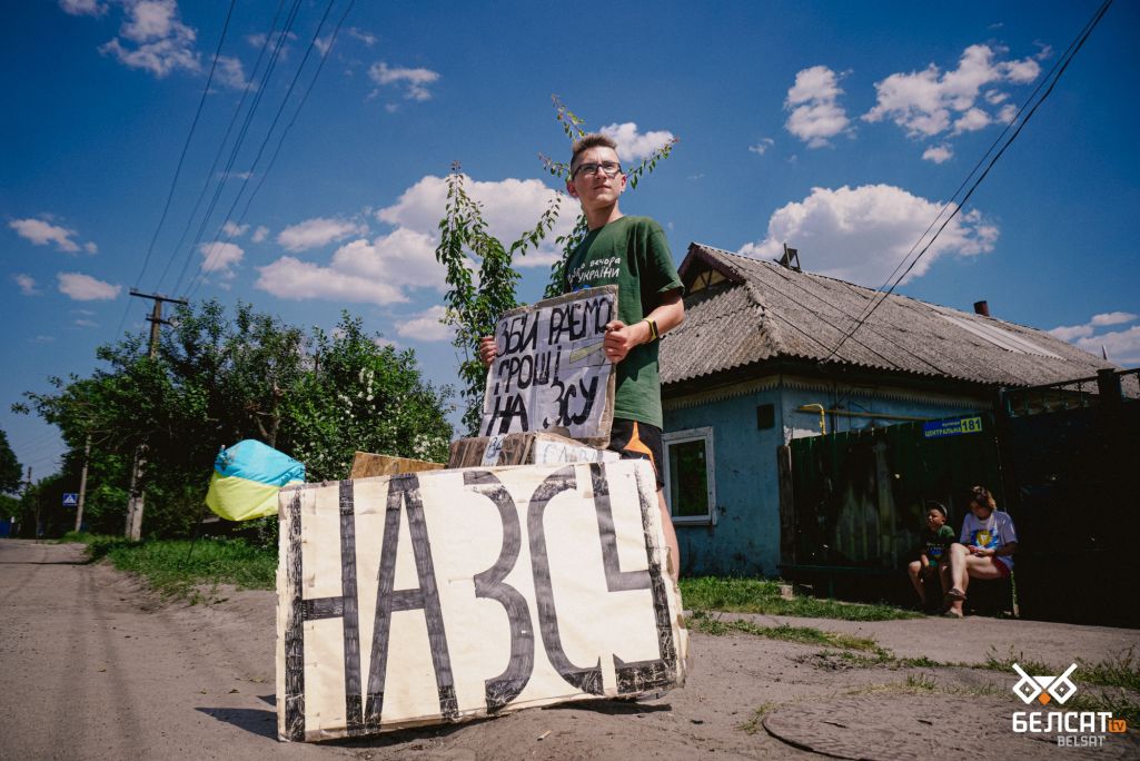 Алег Максюта каля імправізаванага пункту збору фінансавай дапамогі для Збройных сілаў Украіны. Хлопчык падмяняе малодшага брата, пакуль той адпачывае ад спякоты. (Фота: Алесь Усцінаў / Белсат)