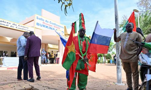 Mężczyzna w tradycyjnym ludowym stroju z rosyjską i burkińską flagą przed salą konferencyjną, w której odbywały rozmowy narodowe w sprawie wyznaczenia tymczasowego prezydenta Burkiny Faso 14 października 2022 r. Fot. ANNE MIMAULT / Reuters / Forum